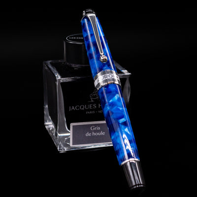 Aurora Optima Auroloide Blue & Chrome fountain pen shown capped, resting at an angle against an ink bottle. The deep marbled blue resin barrel features rhodium-plated trim and the signature Aurora cap band engraving.