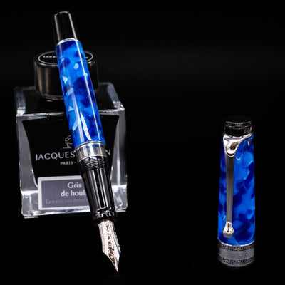 Aurora Optima Auroloide Blue & Chrome fountain pen displayed uncapped, leaning on an ink bottle to reveal its rhodium-plated 14k gold nib. The marbled blue resin barrel is complemented by a matching cap and polished silver-tone accents.