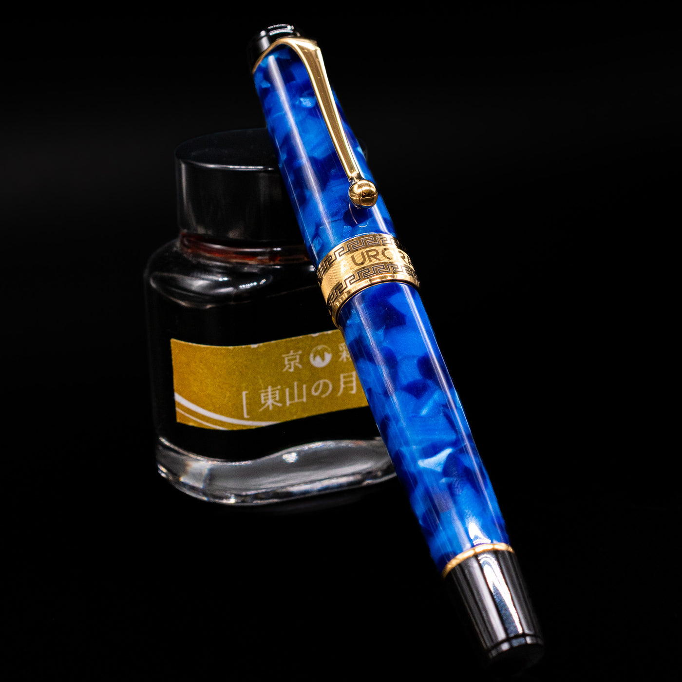 Aurora Optima Auroloide Blue & Gold fountain pen shown capped, resting at an angle against an ink bottle. The deep marbled blue resin barrel features rich gold-plated trim and the signature Aurora cap band engraving.