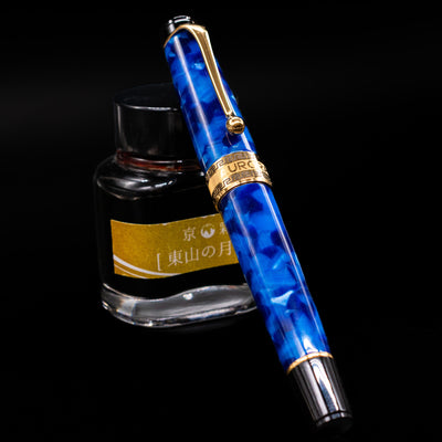 Aurora Optima Auroloide Blue & Gold fountain pen shown capped, resting at an angle against an ink bottle. The deep marbled blue resin barrel features rich gold-plated trim and the signature Aurora cap band engraving.