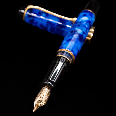 Aurora Optima Auroloide Blue & Gold fountain pen and cap crossed, with focus on the engraved gold nib and the vibrant marbled blue resin.