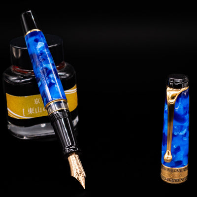 Aurora Optima Auroloide Blue & Gold fountain pen displayed uncapped, leaning on an ink bottle to reveal its gold 14k nib. The marbled blue resin barrel is complemented by a matching cap and polished gold accents.
