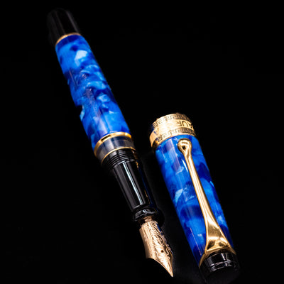 Aurora Optima Auroloide Blue & Gold fountain pen displayed uncapped, revealing the marbled blue resin barrel, gold-plated grip section, and engraved gold nib.