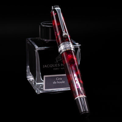 Aurora Optima Auroloide Burgundy fountain pen shown capped, resting at an angle against an ink bottle. The deep marbled burgundy resin barrel features gold-plated trim and the signature Aurora cap band engraving.