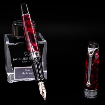 Aurora Optima Auroloide Burgundy fountain pen displayed uncapped, leaning on an ink bottle to reveal its rhodium-plated 14k nib. The rich marbled burgundy resin barrel is complemented by a matching cap and silver accents.