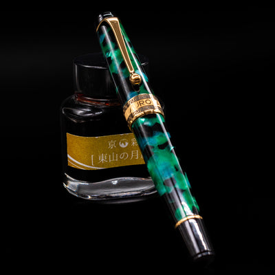 Aurora Optima Auroloide Emerald Green fountain pen shown capped, resting at an angle against an ink bottle. The vibrant marbled green resin barrel features gold-plated trim and the signature Aurora cap band engraving.