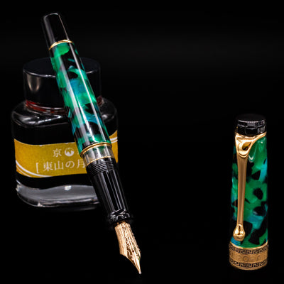 Aurora Optima Auroloide Emerald Green fountain pen displayed uncapped, leaning on an ink bottle to reveal its gold 14k nib. The rich marbled green resin barrel is complemented by a matching cap and warm gold accents.