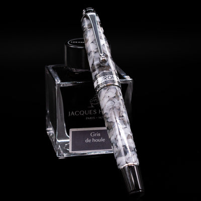Aurora Optima Auroloide Nero Perla fountain pen shown capped, resting at an angle against an ink bottle. The elegant marbled black-and-grey resin barrel features rhodium-plated trim and the signature Aurora cap band engraving.