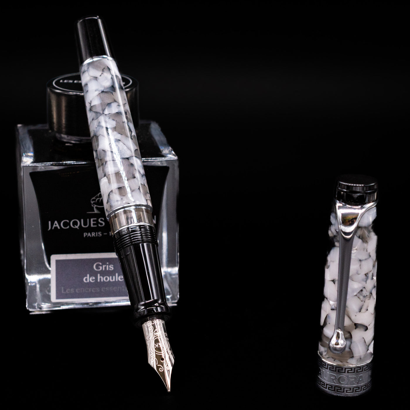 Aurora Optima Auroloide Nero Perla fountain pen displayed uncapped, leaning on an ink bottle to reveal its rhodium-plated 14k gold nib. The smoky marbled resin barrel is complemented by a matching cap and polished silver-tone accents.
