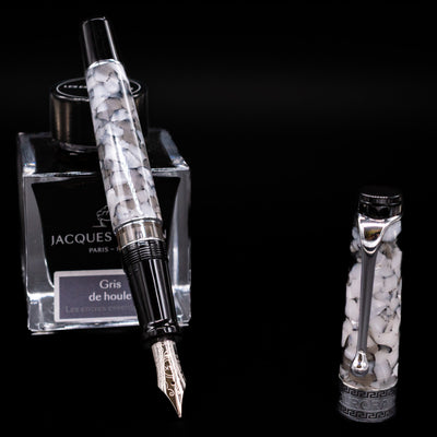 Aurora Optima Auroloide Nero Perla fountain pen displayed uncapped, leaning on an ink bottle to reveal its rhodium-plated 14k gold nib. The smoky marbled resin barrel is complemented by a matching cap and polished silver-tone accents.