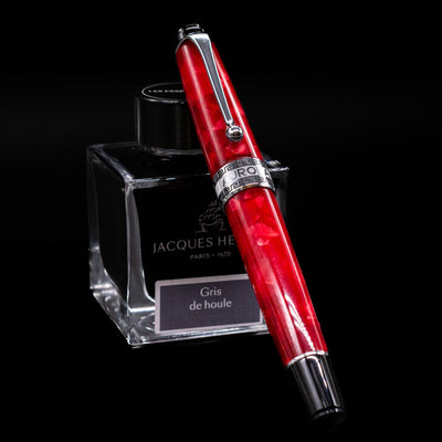 Aurora Optima Auroloide Rossa fountain pen shown capped, resting at an angle against an ink bottle. The vibrant marbled red resin barrel features rhodium-plated trim and the signature Aurora cap band engraving.
