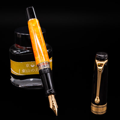 Aurora Optima O Sole Mio fountain pen displayed uncapped, leaning on an ink bottle to reveal its gold 14k nib. The deep black resin cap and section contrast with the bright marbled orange barrel and warm gold trim.