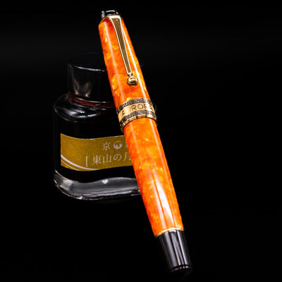 Aurora Optima Orange fountain pen shown capped, resting at an angle against an ink bottle. The vibrant orange resin barrel features a marbled effect with gold-plated trim and the classic Aurora cap band engraving.