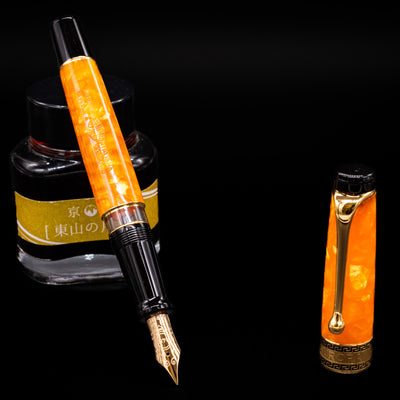Aurora Optima Orange fountain pen displayed uncapped, leaning on an ink bottle to reveal its gold-plated 14k nib. The marbled orange resin barrel contrasts beautifully with the warm gold trim.