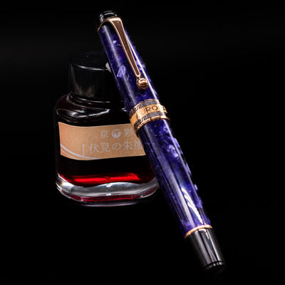 Aurora Optima Viola fountain pen shown capped, resting at an angle against an ink bottle. The rich purple resin barrel features a marbled effect with rhodium-plated trim and the classic Aurora cap band engraving.
