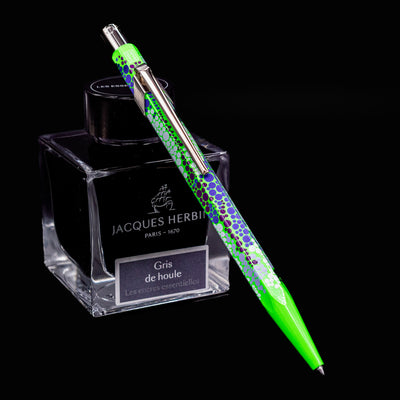 Caran d’Ache 849 Special Edition Germanier Fluo Green ballpoint pen resting at an angle against an ink bottle. The pen features a bright fluorescent green hexagonal aluminum body with a sleek silver clip and push button.