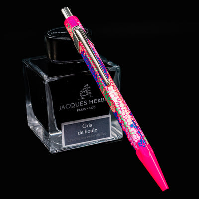 Caran d’Ache 849 Special Edition Germanier Fluo Pink ballpoint pen resting at an angle against an ink bottle. The pen features a vibrant fluorescent pink hexagonal aluminum body with a sleek silver clip and push button.