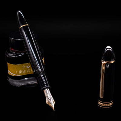 Preowned Montblanc Meisterstück 149 fountain pen displayed uncapped, leaning on an ink bottle to reveal its oversized 18k bi-color gold nib. The deep black resin barrel is accented with gold bands and a sturdy gold clip.