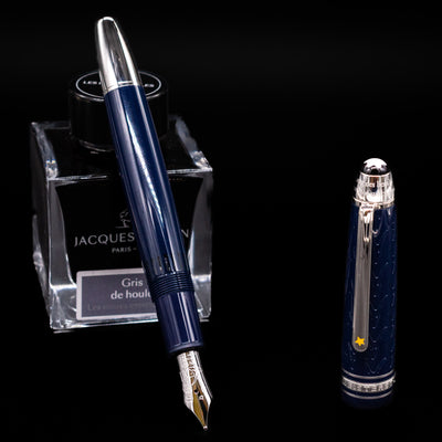 Preowned Montblanc Meisterstück Le Petit Prince Fox LeGrand fountain pen displayed uncapped, leaning on an ink bottle to reveal its rhodium-coated 14k gold nib engraved with the fox motif. The blue resin barrel and platinum accents give it a refined look.