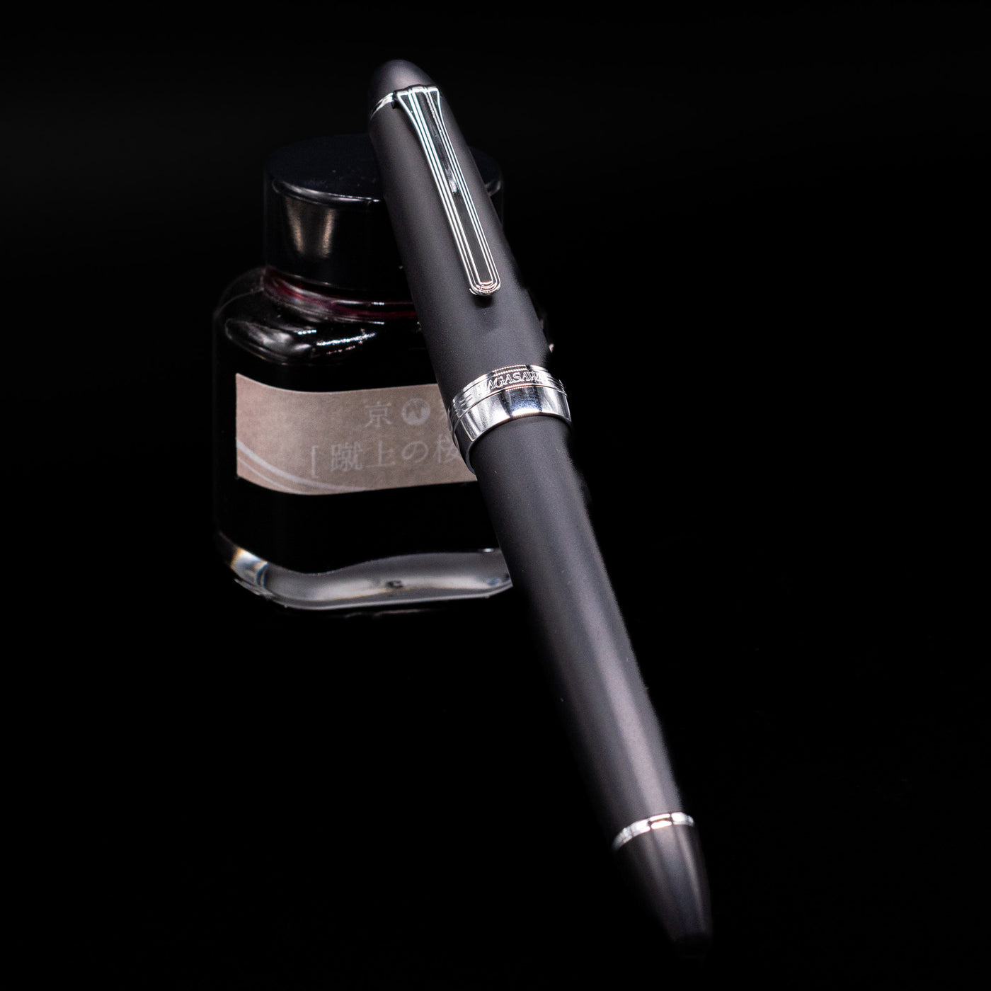 Nagasawa Original Large Fountain Pen - Black & Silver FL (For Lawyer) capped, leaning against an ink bottle. 