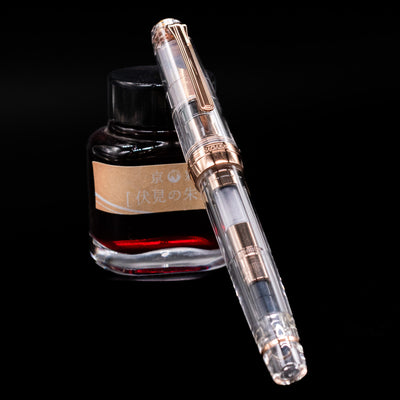 Nagasawa Original Professional Gear Fountain Pen - Skeleton Pink Goldc apped, leaning against an ink bottle. 