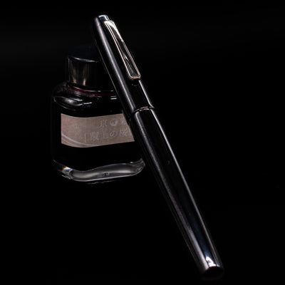 Nakaya Piccola Long Fountain Pen - Kuro Tame capped, leaning against an ink bottle. 