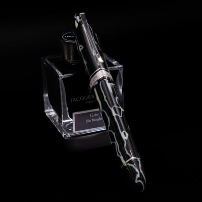 Preowned OMAS 360 Wild Celluloid fountain pen shown capped, resting at an angle against an ink bottle. The triangular-shaped barrel features a striking mix of black, amber, and cream celluloid patterns, complemented by gold-plated trim.