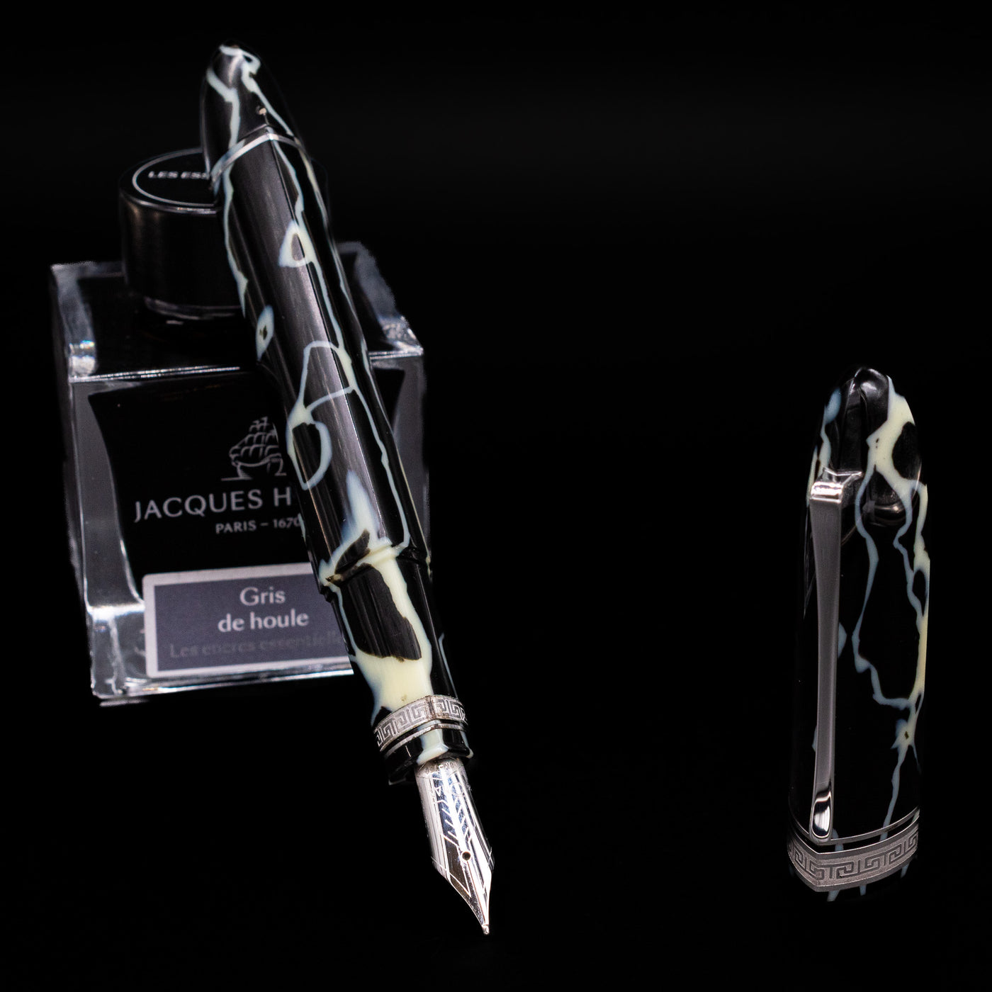 Preowned OMAS 360 Wild Celluloid fountain pen displayed uncapped, leaning on an ink bottle to reveal its 18k gold nib. The pen’s distinctive triangular body showcases swirling black, amber, and cream celluloid with silver accents.