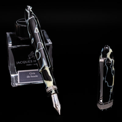 Preowned OMAS 360 Wild Celluloid fountain pen displayed uncapped, leaning on an ink bottle to reveal its 18k gold nib. The pen’s distinctive triangular body showcases swirling black, amber, and cream celluloid with silver accents.