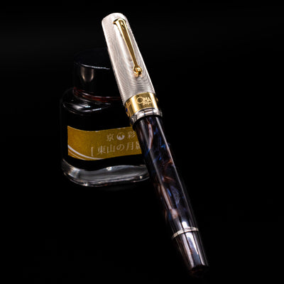 OMAS Bologna Fountain Pen - Galassia Argento capped, leaning against an ink bottle. 