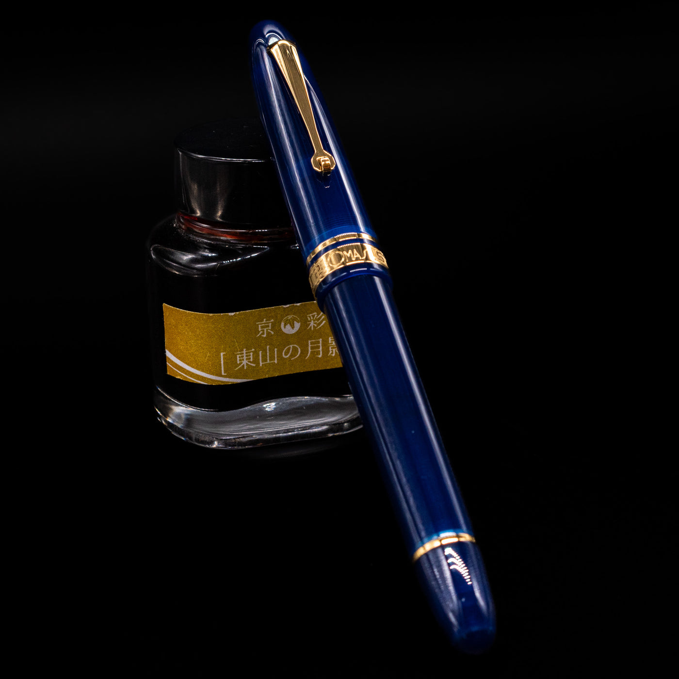 Omas Ogiva Fountain Pen - Blue Gold capped, leaning against an ink bottle. 