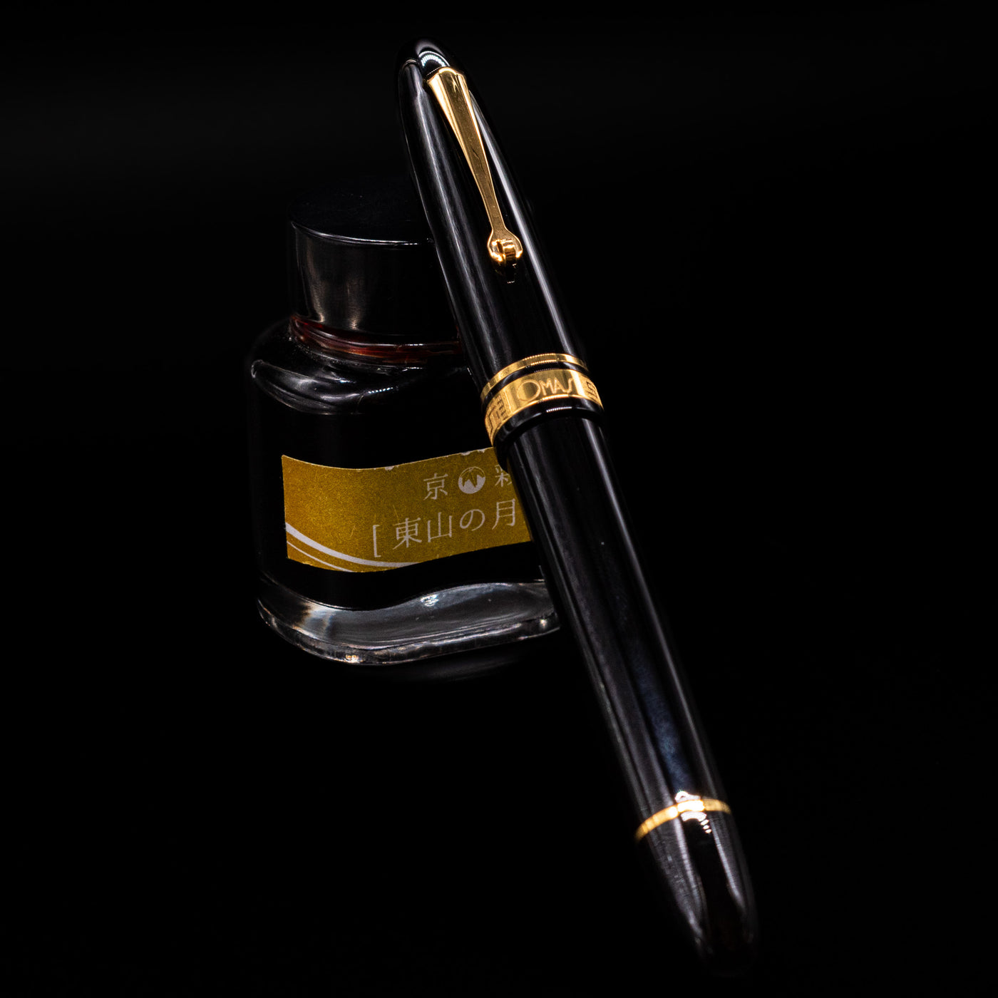 Omas Ogiva Fountain Pen - Nera Gold capped, leaning against an ink bottle. 