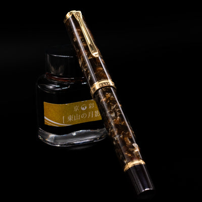 Pelikan Souverän M1000 Renaissance Brown fountain pen shown capped, resting against an ink bottle. The marbled brown resin barrel swirls with deep and warm tones, accented by glossy black resin and gold-plated trim.