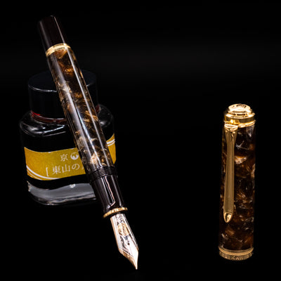 Pelikan Souverän M1000 Renaissance Brown fountain pen displayed uncapped, leaning on an ink bottle to reveal its large 18k bi-color gold nib. The rich marbled brown barrel contrasts with the glossy black cap and gold details.