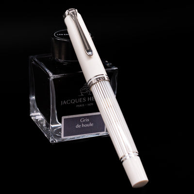 Pelikan Souverän M605 White Transparent fountain pen shown capped, resting at an angle against an ink bottle. The glossy white resin body features translucent barrel stripes with elegant palladium-plated trim.