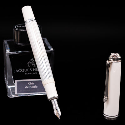 Pelikan Souverän M605 White Transparent fountain pen displayed uncapped, leaning on an ink bottle to reveal its rhodium-plated 14k gold nib. The translucent striped barrel and bright white cap are accented with palladium trim.