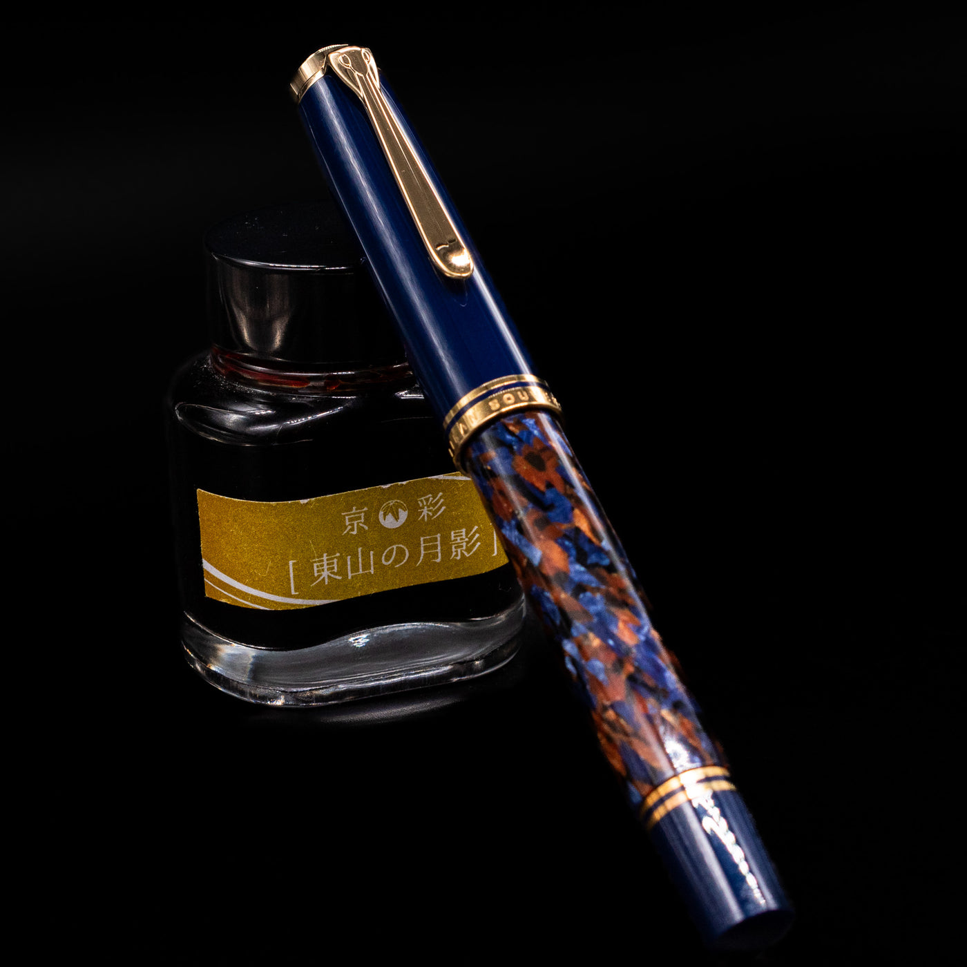 Preowned Pelikan Souverän M800 Stone Garden fountain pen shown capped, resting at an angle against an ink bottle. The marbled deep blue resin barrel is paired with a dark blue cap and gold-plated trim for a rich, sophisticated look.