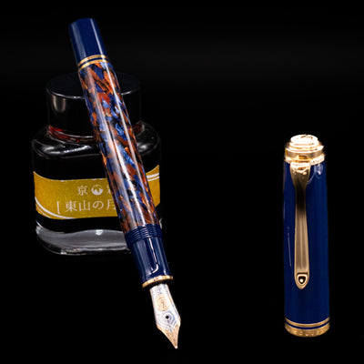 Preowned Pelikan Souverän M800 Stone Garden fountain pen displayed uncapped, leaning on an ink bottle to reveal its bi-color 18k gold nib. The marbled blue barrel and dark blue cap are accented with gleaming gold trim.