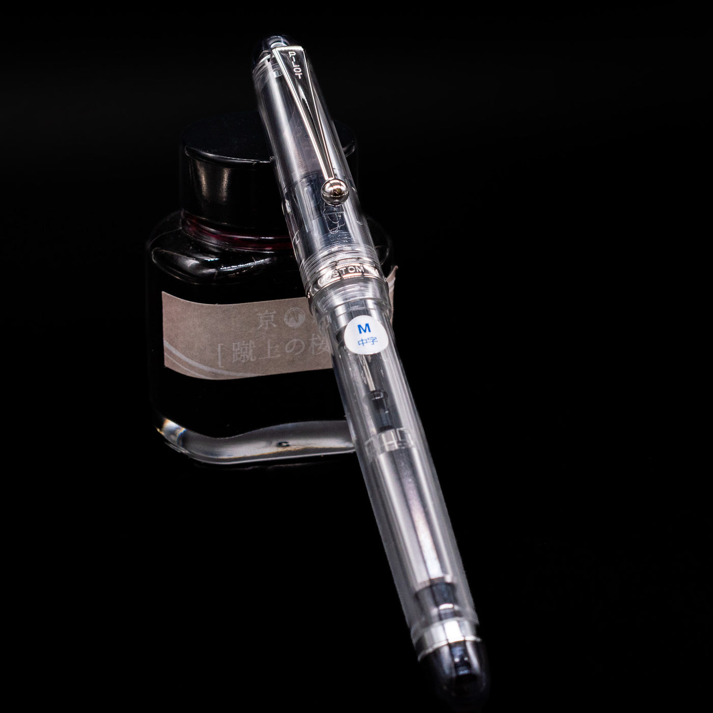 Pilot Custom 74 Fountain Pen - Clear capped, leaning against an ink bottle. 
