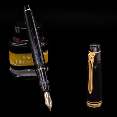 Preowned Pilot Justus 95 Net Black fountain pen displayed uncapped, leaning on an ink bottle to reveal its 14k gold adjustable nib. The pen’s black net-pattern barrel and gold accents are highlighted in soft lighting.