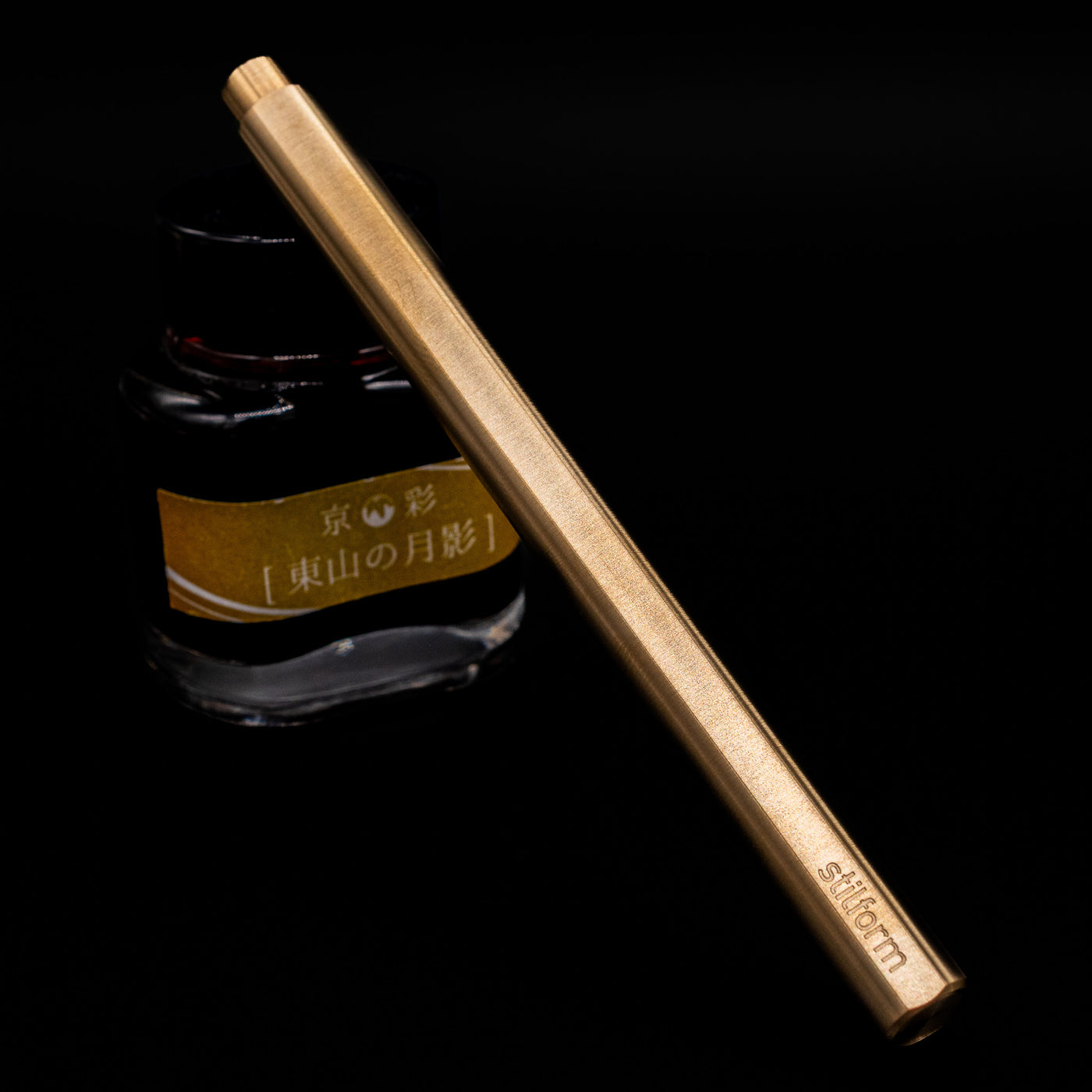 Stilform FLOW Rollerball Pen – Brass Radial Brushed capped, leaning against an ink bottle. 
