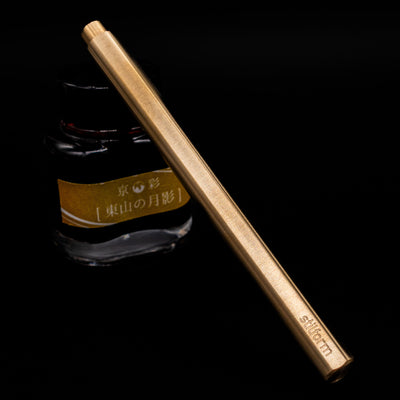 Stilform FLOW Rollerball Pen – Brass Radial Brushed capped, leaning against an ink bottle. 
