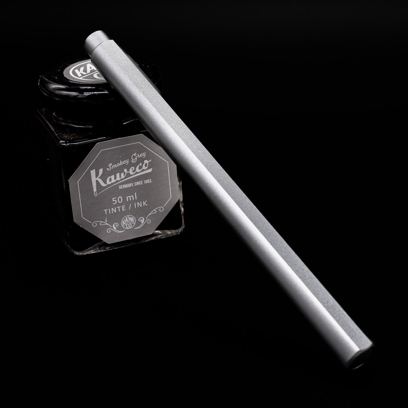 Stilform FLOW Rollerball Pen – Comet Grey Aluminum capped leaning on an ink bottle.