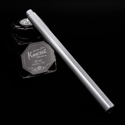 Stilform FLOW Rollerball Pen – Comet Grey Aluminum capped leaning on an ink bottle.