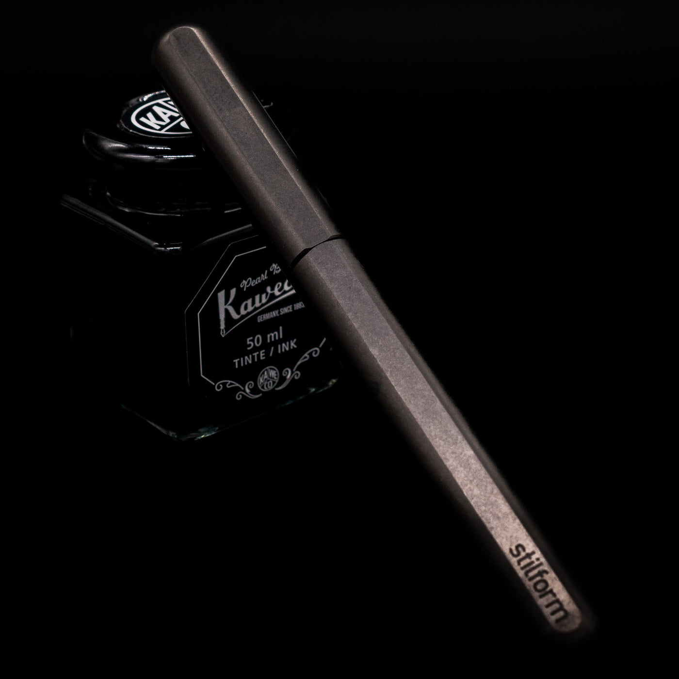 Stilform INK Fountain Pen – Titanium DLC capped, leaning against an ink bottle. 