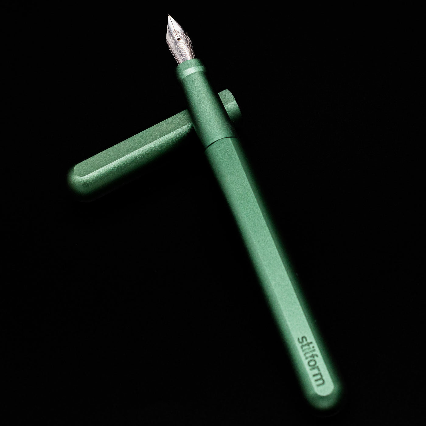 Stilform INK Fountain Pen – Aurora Green Aluminum laying on top of its cap in a crossed position showing full view of the entire uncapped pen.