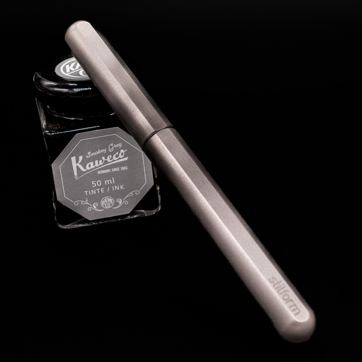 Stilform INK Fountain Pen – Titanium Matte capped, leaning against an ink bottle. 