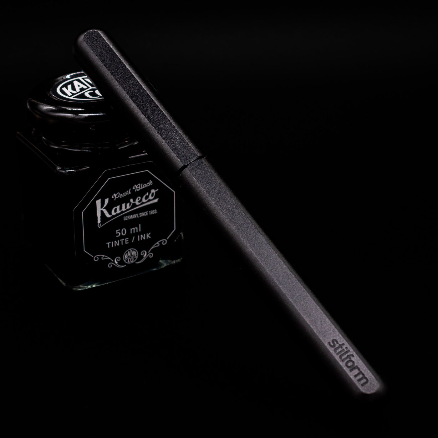 Stilform INK Fountain Pen – Warp Black Aluminum capped, leaning against an ink bottle. 