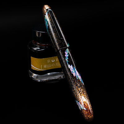 Taccia Miyabi Bon Bori Aurora Glimmer Fountain Pen capped, leaning against an ink bottle. 