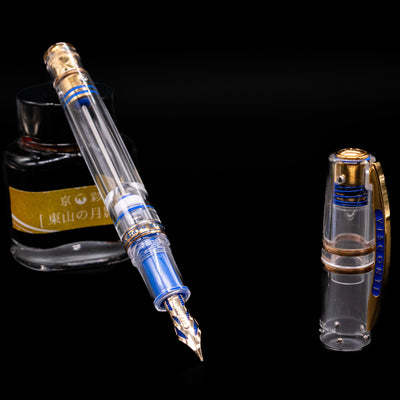 A transparent Visconti Homo Sapiens Full Demo fountain pen, uncapped with its nib exposed, leaning on an ink bottle.